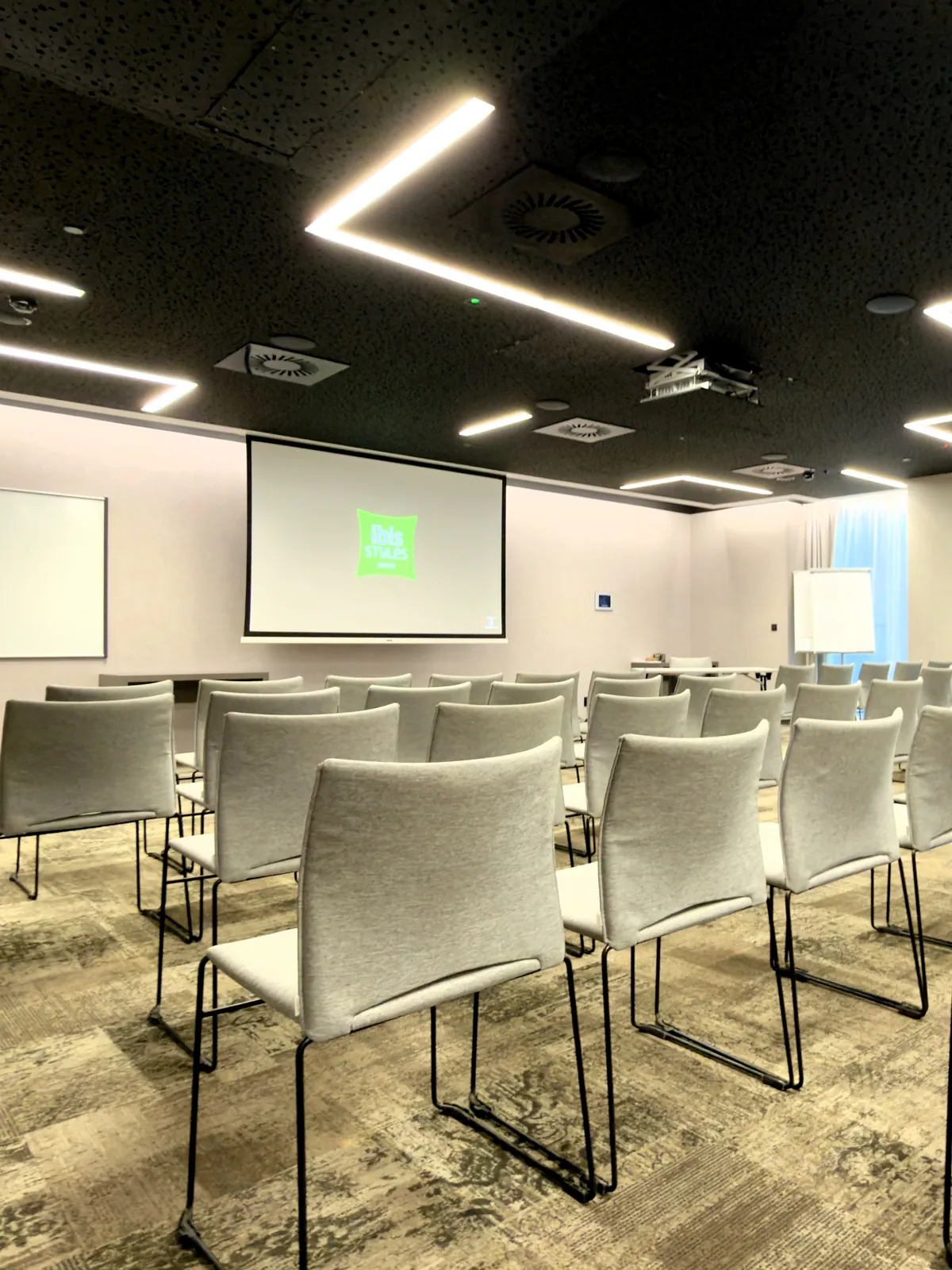 Sala konferencyjna ibis Styles Warszawa City w ustawieniu teatralnym — rzędy krzeseł, ekran projekcyjny z logo ibis Styles, flipchart i okna z naturalnym światłem