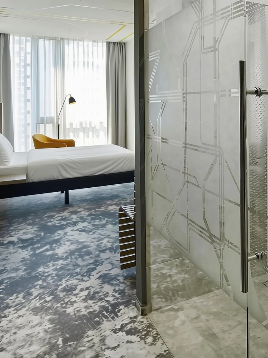 Pokój standardowy w ibis Styles Warszawa City — łóżko, żółty fotel, panoramiczne okno i szklana ścianka łazienki z wytrawioną mapą warszawskich tramwajów