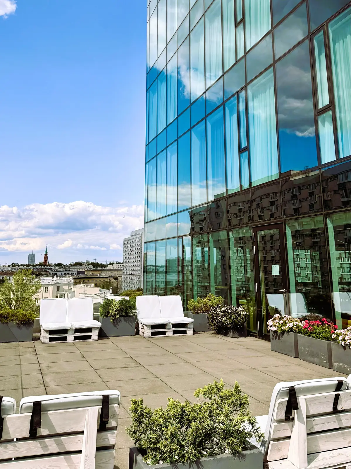 Taras na 7. piętrze — leżaki z palet, donice z kwiatami, szklana fasada hotelu odbijająca chmury i panorama Warszawy w tle